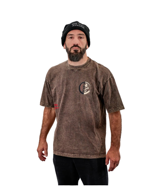 A man wearing a brown acid-wash t-shirt with a circular chest graphic, a black beanie, and black pants, standing against a white background.