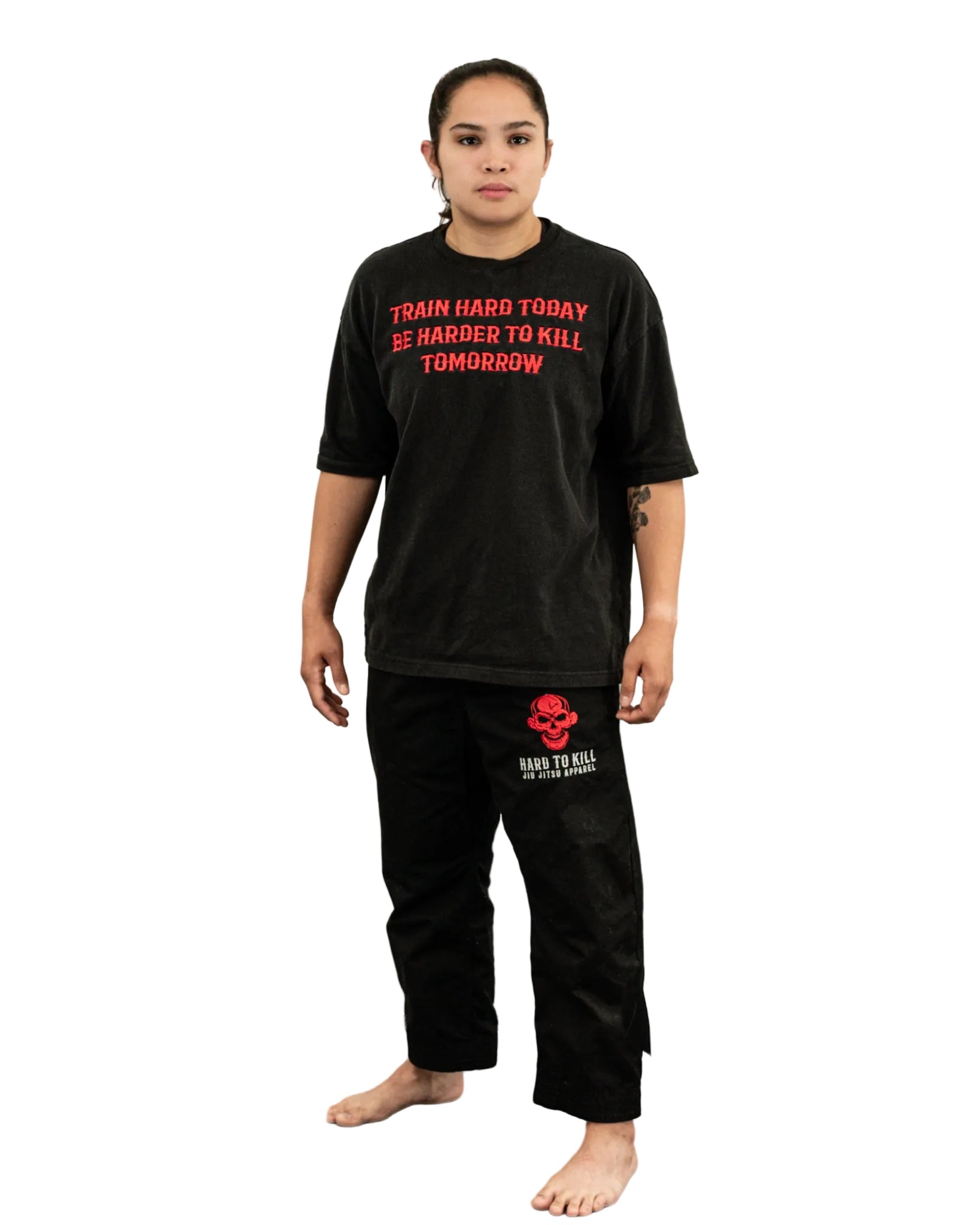 A person standing barefoot in a black martial arts uniform with red printed text on the chest and a red skull logo on the thigh, photographed against a white studio backdrop with a gray floor.