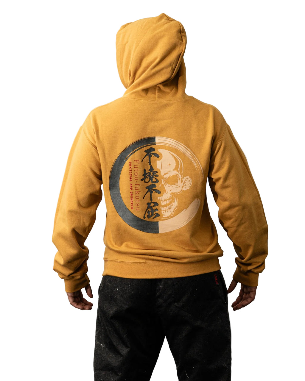 A person shown from the back wearing a mustard yellow hoodie with a large circular graphic print on the back in black, gray, and beige, paired with black pants against a plain white background.